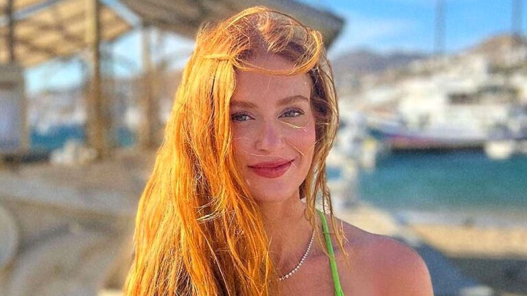 Atriz ruiva posa ao ar livre com cabelos ao vento e paisagem litorânea desfocada ao fundo