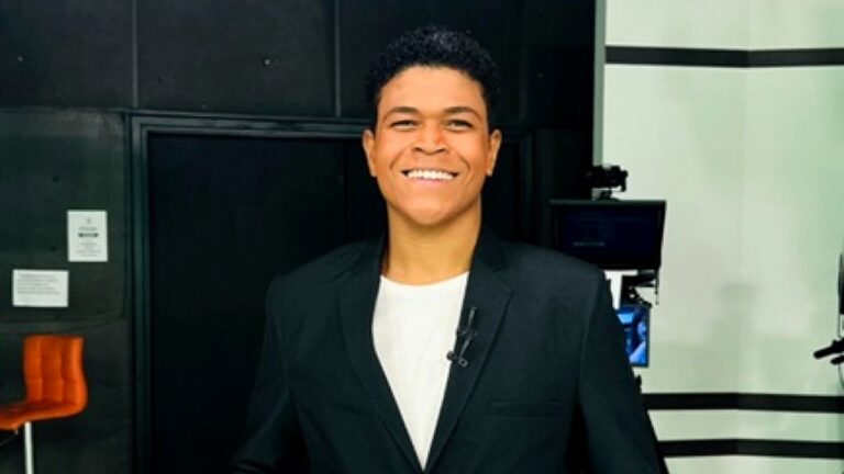 Homem sorri nos bastidores de estúdio de TV usando blazer preto e microfone de lapela