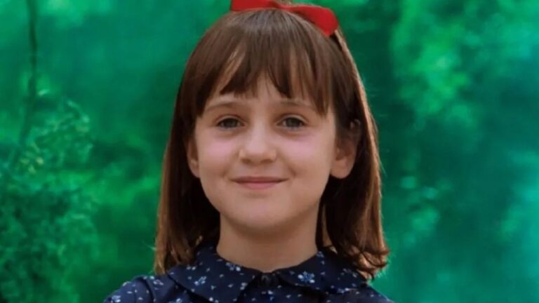 Mara Wilson sorri com laço vermelho no cabelo em cena infantil do filme Matilda diante de fundo verde desfocado