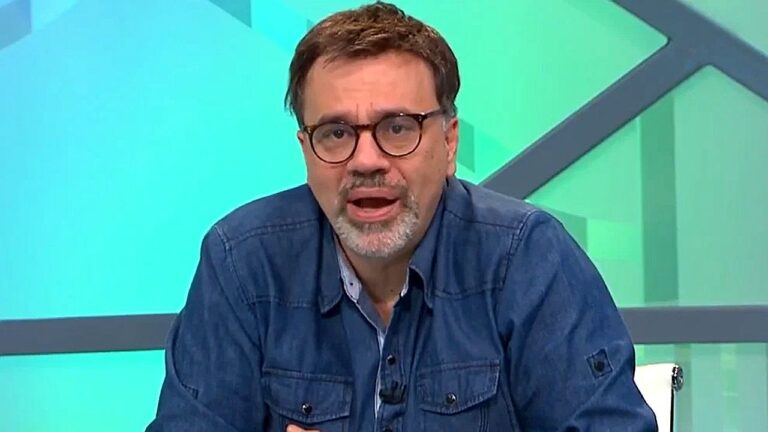 Apresentador de óculos e camisa jeans fala no estúdio de programa de TV com cenário em tons de verde