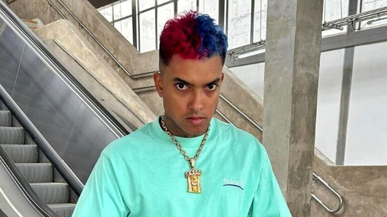 Homem com cabelo metade vermelho e metade azul posa em escada rolante usando camiseta verde e corrente no pescoço