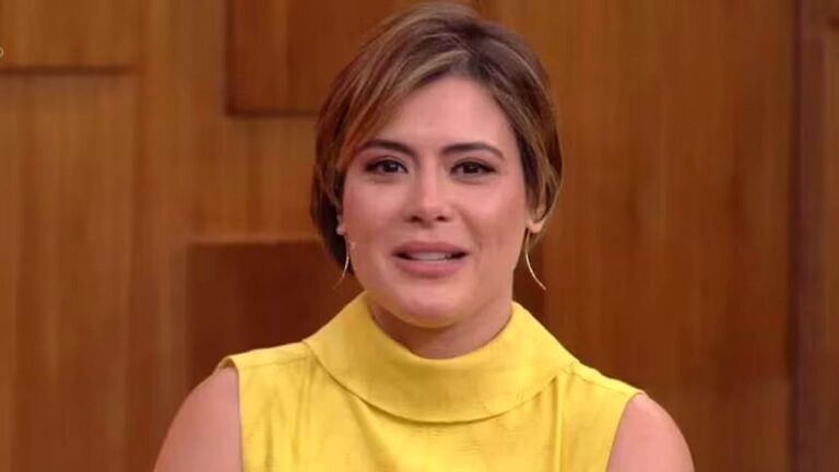 Mulher de vestido amarelo fala em programa de TV diante de painel de madeira ao fundo