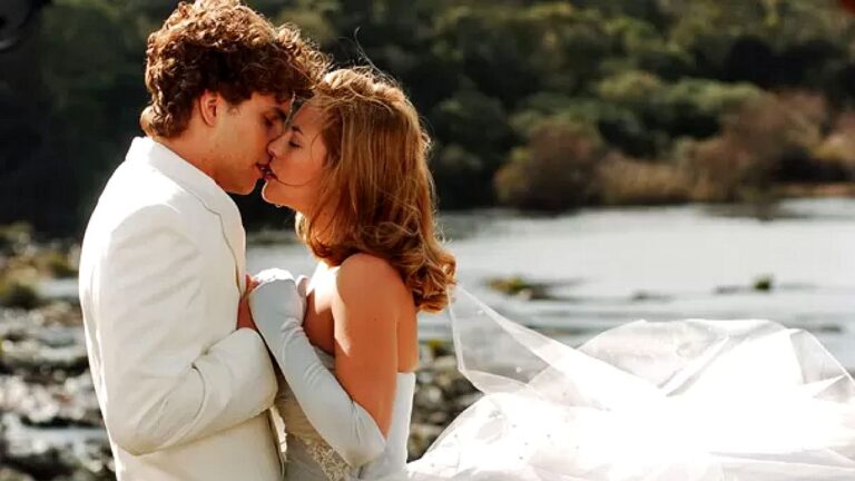Casal vestido de noivos se beija à beira de lago em cena romântica de filme adolescente