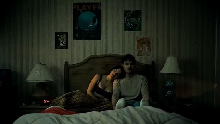 Casal sentado na cama em quarto escuro em cena do filme Obsessão