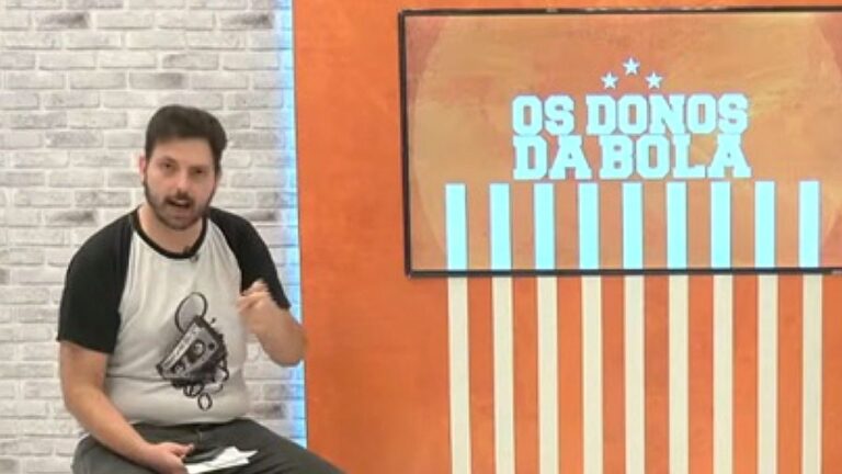 Apresentador comenta futebol ao lado de tela com logotipo do programa Os Donos da Bola