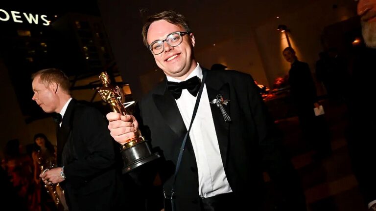 Homem de smoking segura estatueta do Oscar sorrindo em evento noturno com convidados ao fundo