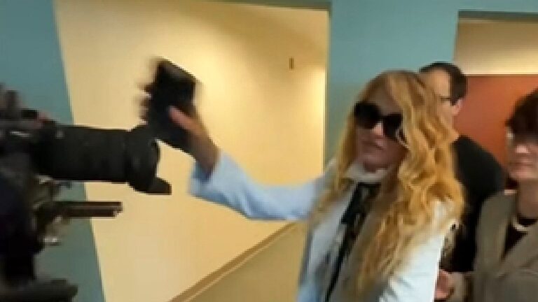 Mulher loira de óculos escuros segura o celular diante de câmera durante gravação em estúdio de TV