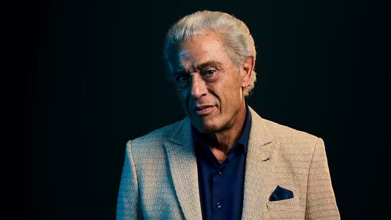 Paulo Gorgulho com blazer claro e camisa azul em retrato promocional com fundo escuro e iluminação dramática