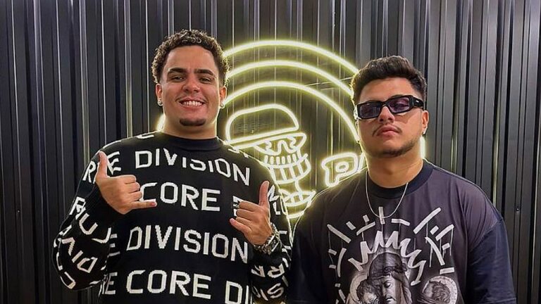 Dois homens posam diante de parede preta com letreiro iluminado ao fundo