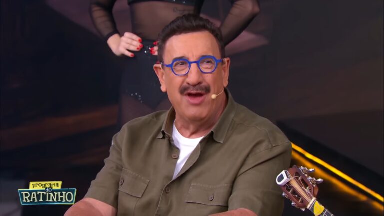 Ratinho fala durante o Programa do Ratinho no SBT com microfone e músico ao fundo em estúdio.