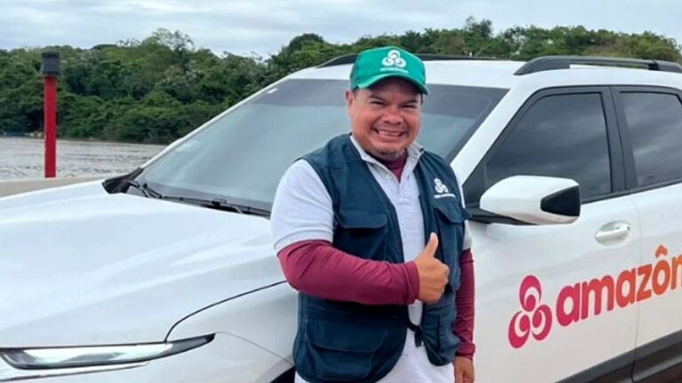 Homem posa ao lado de carro da Rede Amazônica em área ribeirinha usando boné verde e colete da empresa