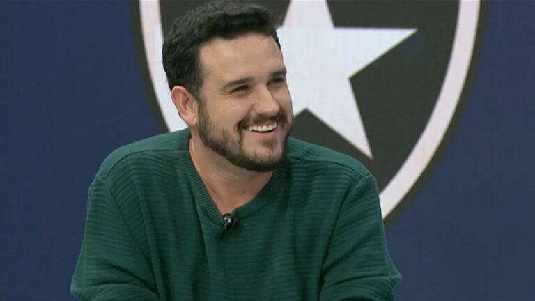 Homem sorri durante participação em programa esportivo com escudo do Botafogo ao fundo