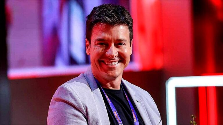Rodrigo Faro sorri durante evento com iluminação vermelha e crachá no pescoço
