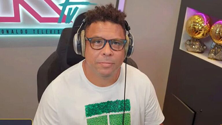 Homem de óculos e fones de ouvido participa de transmissão esportiva em estúdio com troféus ao fundo
