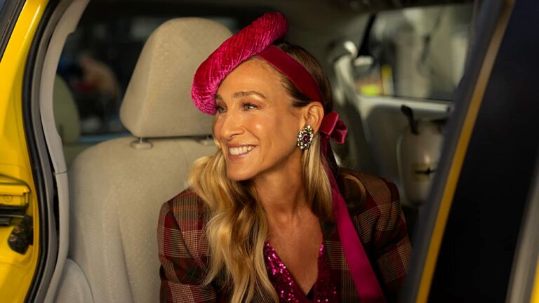 Sarah Jessica Parker sorri ao sair de táxi usando chapéu rosa e roupa brilhante em cena de And Just Like That…