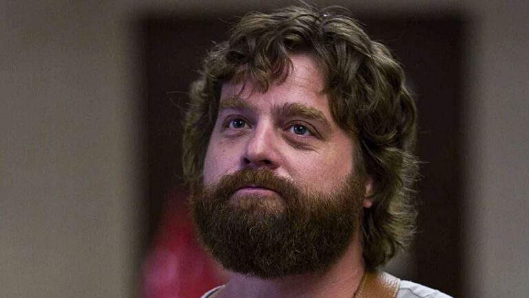 Homem de barba longa e cabelo cacheado observa alguém em cena de série de comédia dramática
