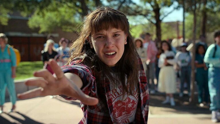 Millie Bobby Brown estende a mão com expressão intensa em cena da série Stranger Things cercada por estudantes