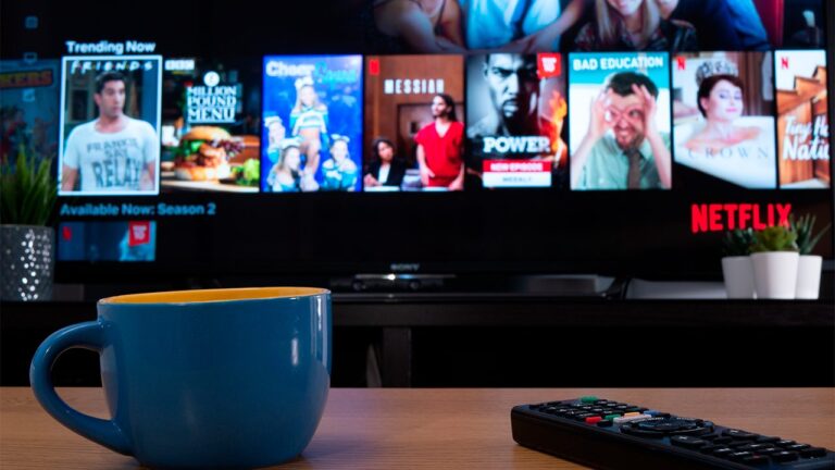 Tela da Netflix exibindo séries e filmes em smart TV com controle remoto e caneca sobre mesa de madeira.