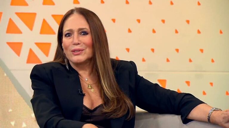 Atriz conversa no sofá de programa de TV usando blazer preto e maquiagem marcante