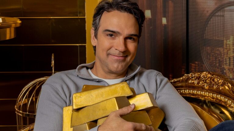 Tadeu Schmidt sorrindo enquanto segura barras de ouro em ambiente luxuoso com decoração dourada ao fundo