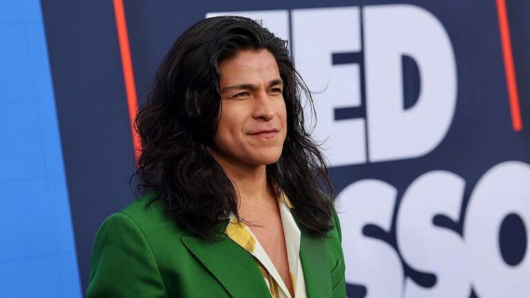 Homem de cabelo comprido posa diante de painel promocional em evento de série de TV