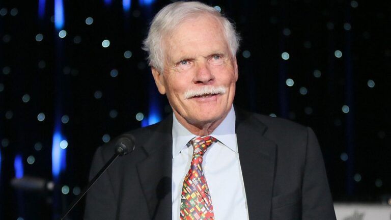 Ted Turner, homm de terno e gravata com bandeiras, discursa em evento com microfone diante de fundo escuro iluminado.