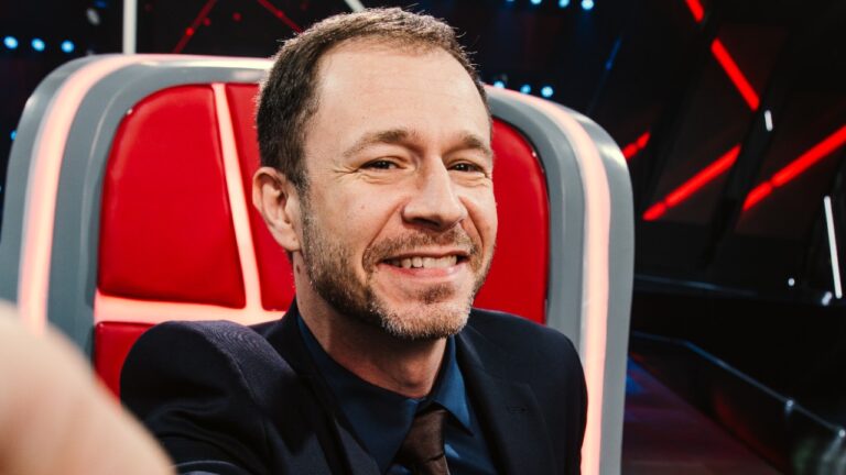 Tiago Leifert sorri sentado na cadeira vermelha do The Voice em estúdio iluminado