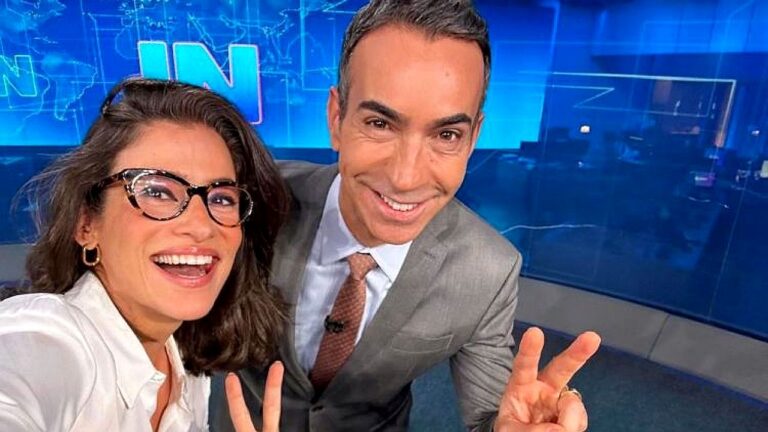 Dois apresentadores sorriem e fazem sinal de paz em selfie no estúdio de telejornal com fundo azul