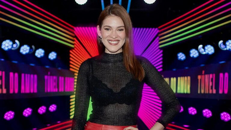 Mulher ruiva posa sorrindo em cenário colorido com luzes neon do programa Túnel do Amor