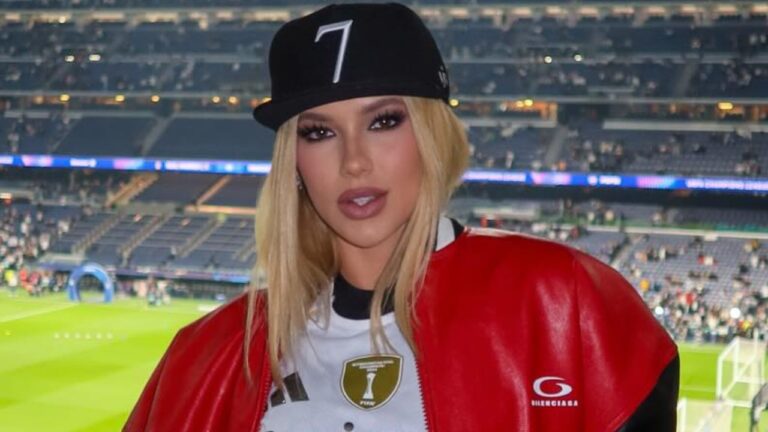 Virginia Fonseca posa em estádio de futebol usando boné preto, jaqueta vermelha e camisa esportiva branca com gramado ao fundo