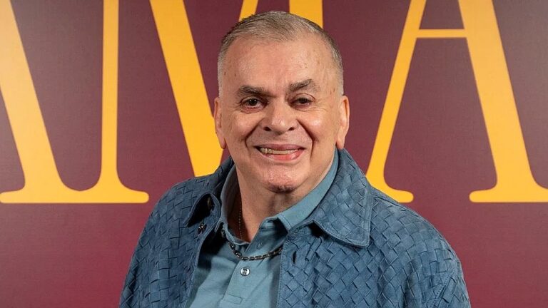 Homem de camisa azul posa sorrindo diante de painel com letras douradas ao fundo