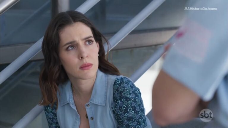Joana, com expressão preocupada e camisa azul, observa alguém à sua frente em cena de A História de Joana, a Virgem