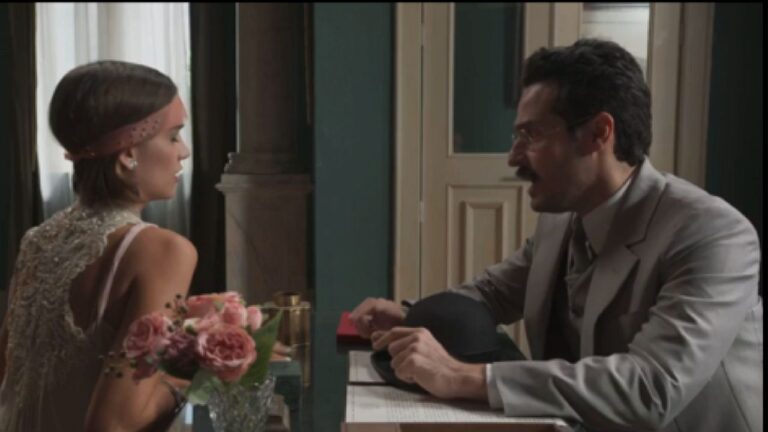 Mulher com vestido elegante e arranjo no cabelo conversa com homem de terno e bigode em cena de novela de época junto a mesa com flores rosas