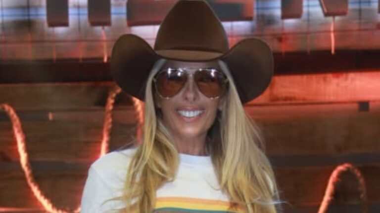 Adriane Galisteu de chapéu cowboy e óculos escuros sorri diante de iluminação decorativa em ambiente interno