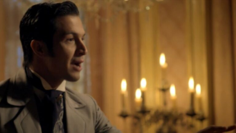 Homem de terno claro e gravata fala em ambiente iluminado por candelabros em cena de novela de época