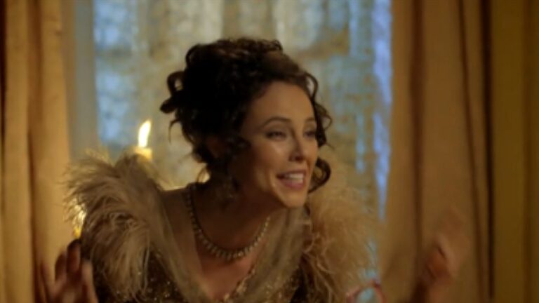 Mulher com penteado elaborado e vestido de época com detalhes de plumas sorri enquanto conversa em salão iluminado por velas em cena de novela histórica