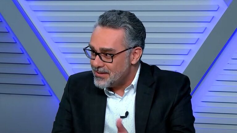 André Marsiglia fala durante programa de debates usando terno preto e óculos em estúdio de televisão