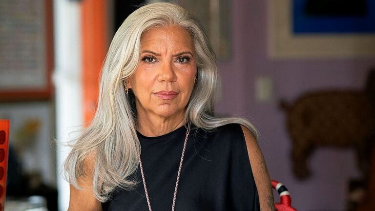 Mulher de cabelos longos grisalhos encara a câmera com expressão firme usando blusa preta sem mangas em ambiente residencial com decoração desfocada ao fundo