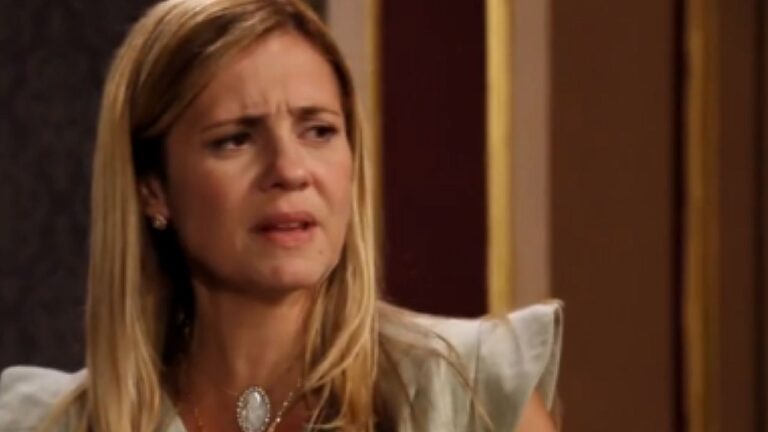 Mulher loira com expressão de preocupação e sobrancelhas franzidas durante conversa em cena de novela ambientada em residência elegante