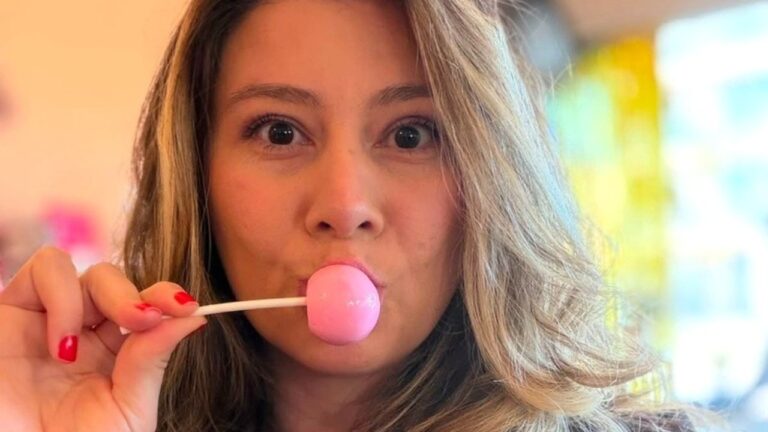 Bella Mantovani segura pirulito rosa próximo à boca e encara a câmera com expressão surpresa em ambiente interno desfocado