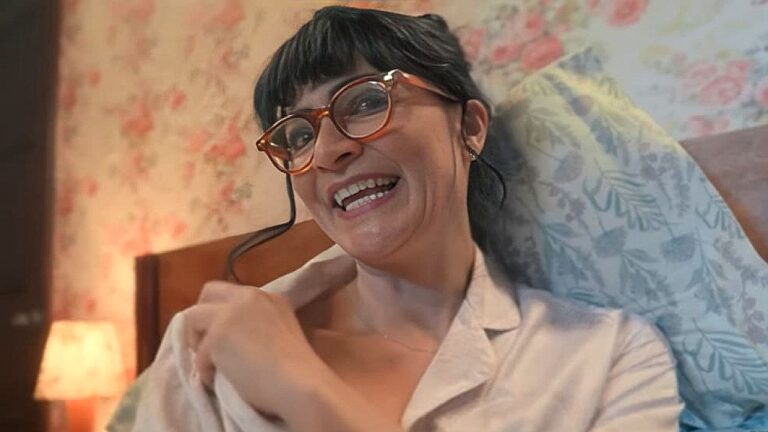 Betty Pinzón sorri usando óculos grandes e pijama em cena da novela Betty, a Feia: A História Continua