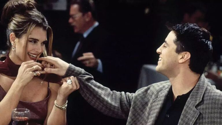 Brooke Shields sorri durante jantar enquanto segura a mão de homem em cena de série ambientada em restaurante elegante