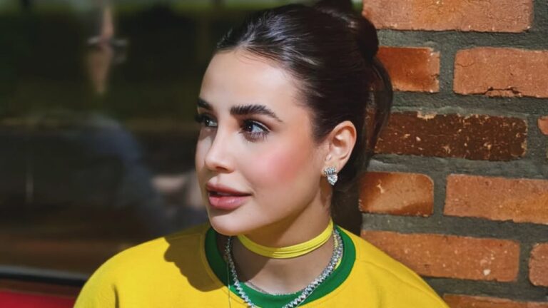 Bruna Pinheiro de blusa amarela e verde olha para o lado diante de parede de tijolos com cabelo preso e colares no pescoço