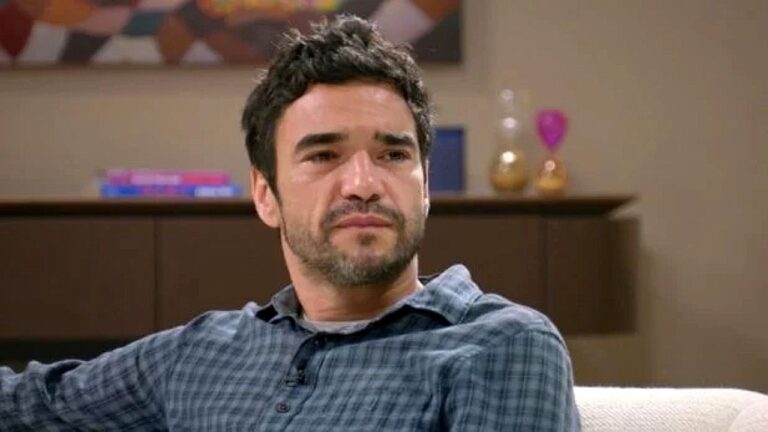 Homem de barba curta e camisa xadrez observa alguém com expressão séria e emocionada em sala de estar com móveis e objetos decorativos ao fundo