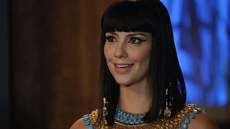 Mulher caracterizada como rainha egípcia com traje dourado e colar azul em cena da novela Os Dez Mandamentos