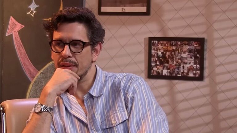 João Vicente de Castro com óculos e mão no queixo durante participação em programa de entrevistas