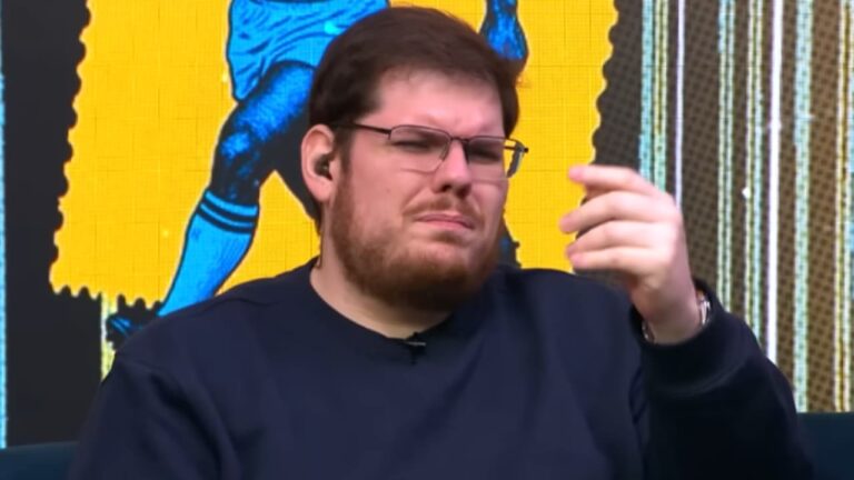 Casimiro Miguel de óculos e moletom escuro faz gesto com a mão durante participação em programa com painel colorido ao fundo