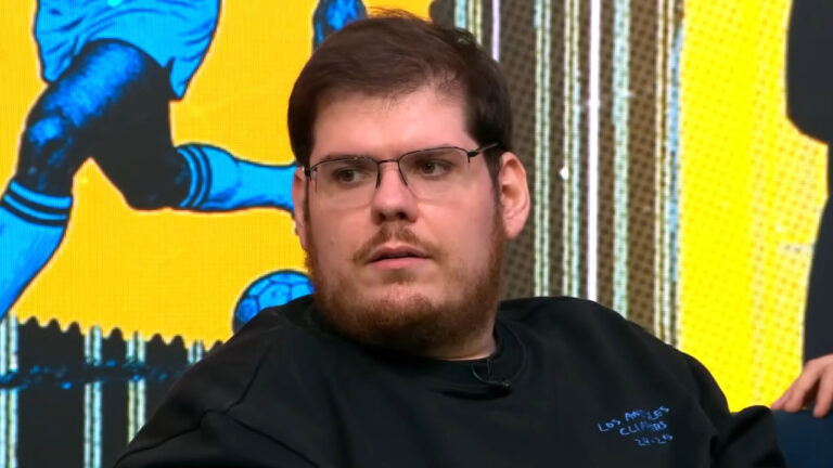 Casimiro Miguel de óculos e camiseta preta durante participação em programa com painel esportivo ao fundo