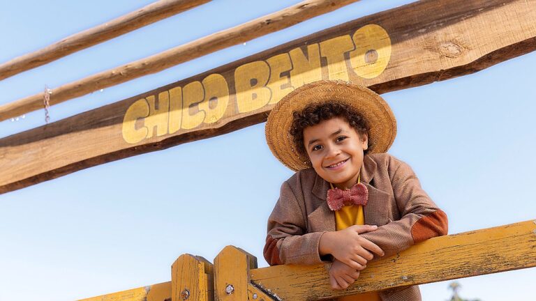 Menino com chapéu de palha sorri apoiado em cerca sob placa Chico Bento em imagem promocional da série infantil