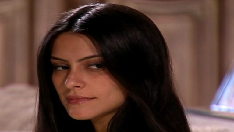Cleo observa alguém de lado com expressão desconfiada e olhar contido em cena de novela ambientada em residência elegante